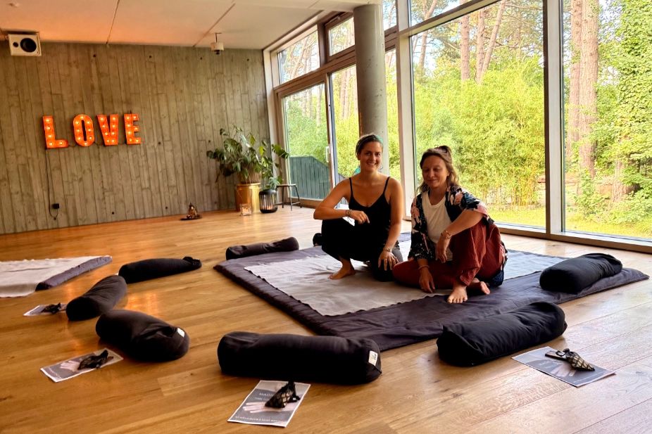 Mirjam Brauer und Mandy Hermann sitzen auf einer Matte in einem Raum mit Bolstern und großen Fenstern